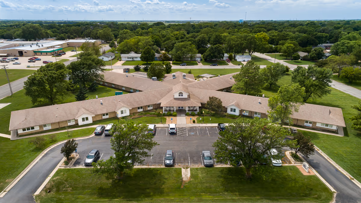 Aerial view of Halstead Health & Rehab facility showing a single-story building with a brown roof and multiple wings extending outward. The building is surrounded by green lawns, trees, and a parking lot with several cars parked. The surrounding area includes residential houses and more greenery under a partly cloudy sky.