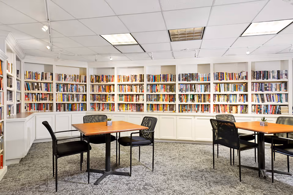 A well-lit library room with white built-in bookshelves filled with books along the walls. There are two wooden tables each surrounded by four black chairs. The floor is carpeted with a patterned design and the ceiling has recessed lighting and track lights.