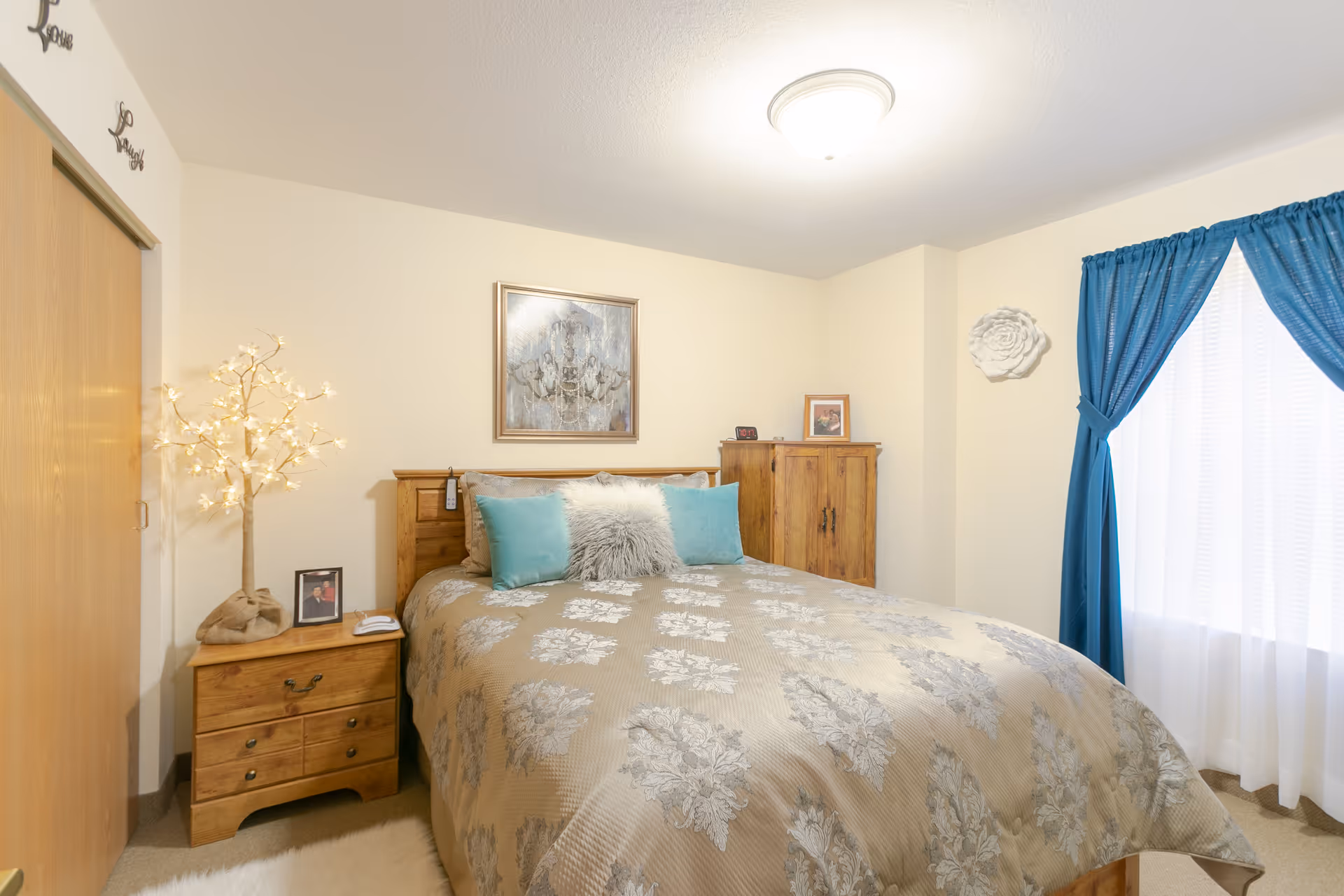 A cozy bedroom with a wooden bed frame and a beige and gray floral patterned comforter. The bed has two teal pillows and a fluffy gray pillow. To the left of the bed is a wooden nightstand with a framed photo, a decorative lighted tree, and a telephone. Above the bed hangs a framed artwork. On the right side of the room is a wooden cabinet and a window with blue curtains tied back, allowing natural light to enter. The walls are painted light beige and the ceiling has a round light fixture.