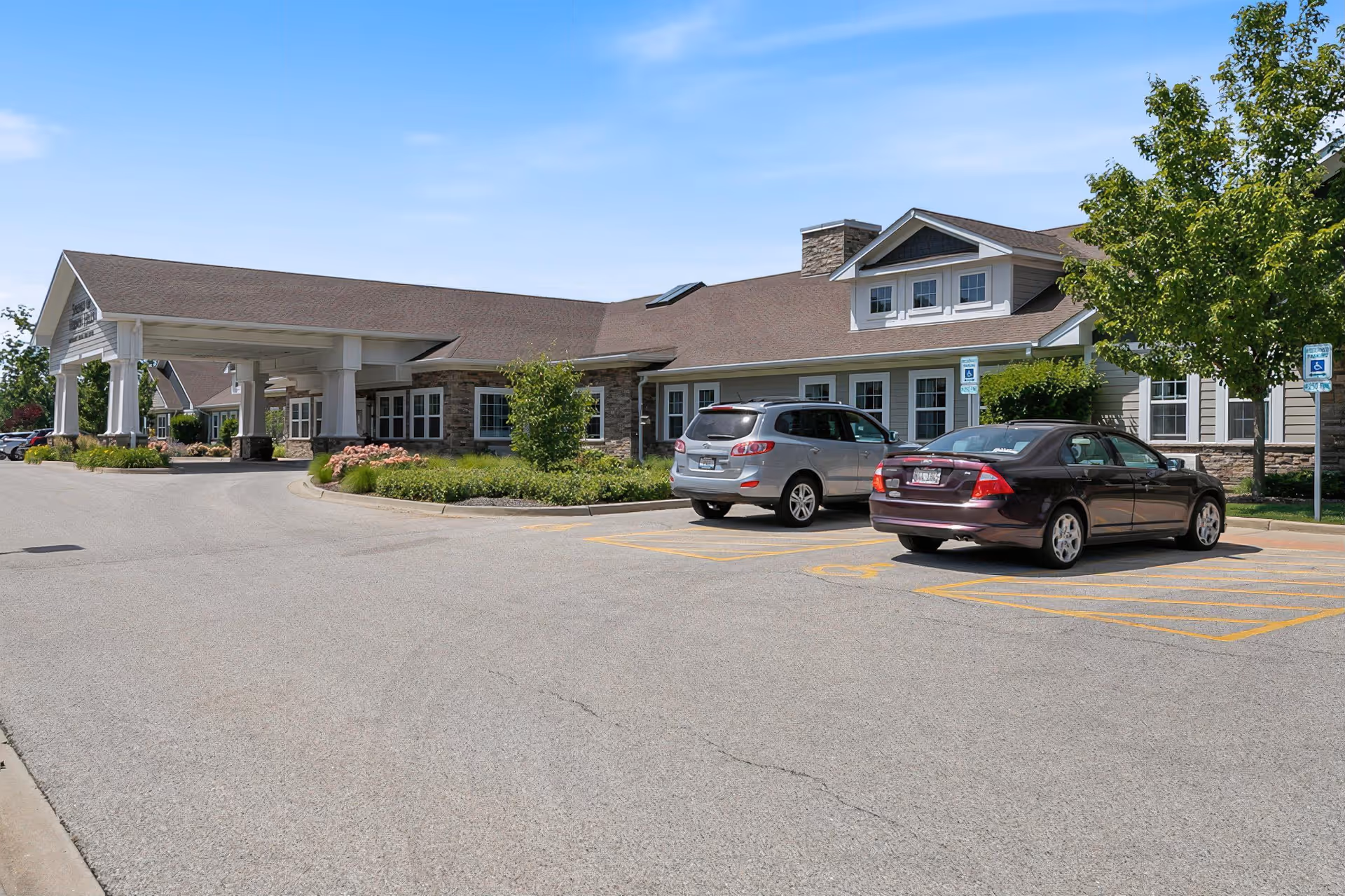 Exterior view of The Laurel at Vernon Hills Memory Care facility showing a single-story building with a covered entrance, several windows, and a parking lot with two cars parked in handicapped spaces. There are trees and landscaping around the building under a clear blue sky.