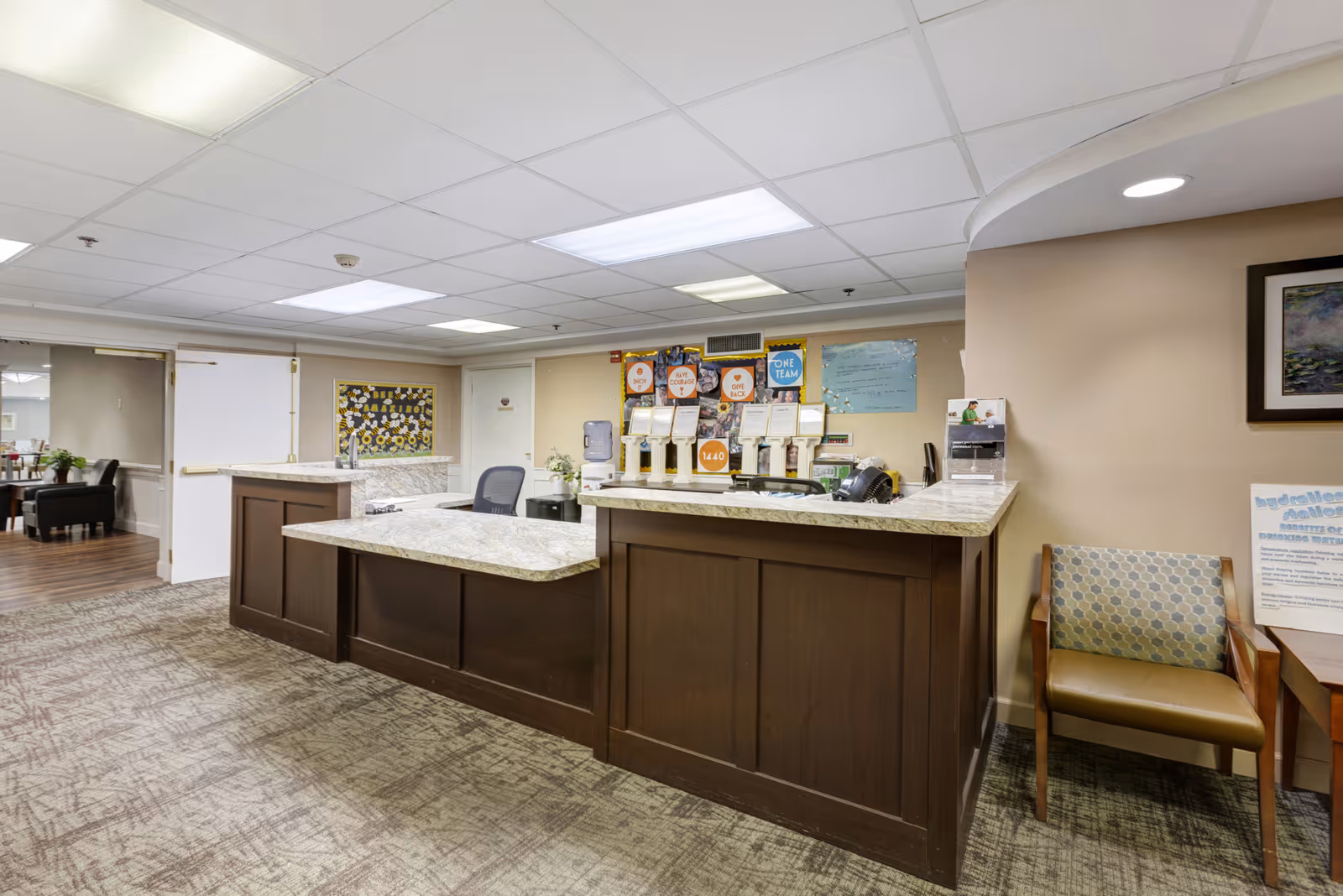 Reception desk area inside a senior living facility with a marble countertop, office chair, informational displays, and a patterned armchair to the right. The room has beige walls, carpeted floor, and fluorescent ceiling lights.