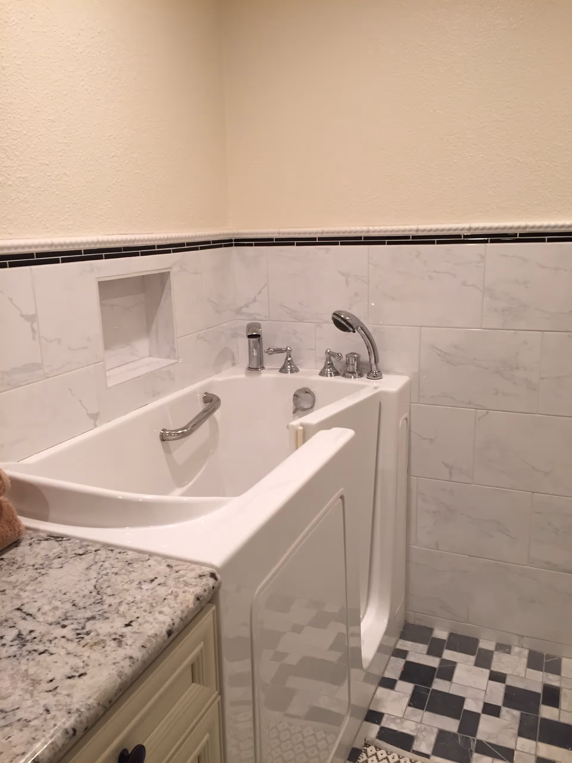 A walk-in bathtub with a side door and chrome fixtures, including a handheld showerhead and faucet, set against a wall with white marble tiles and a black tile border. The floor has a black, white, and gray checkered tile pattern, and a granite countertop with a beige cabinet is partially visible on the left.