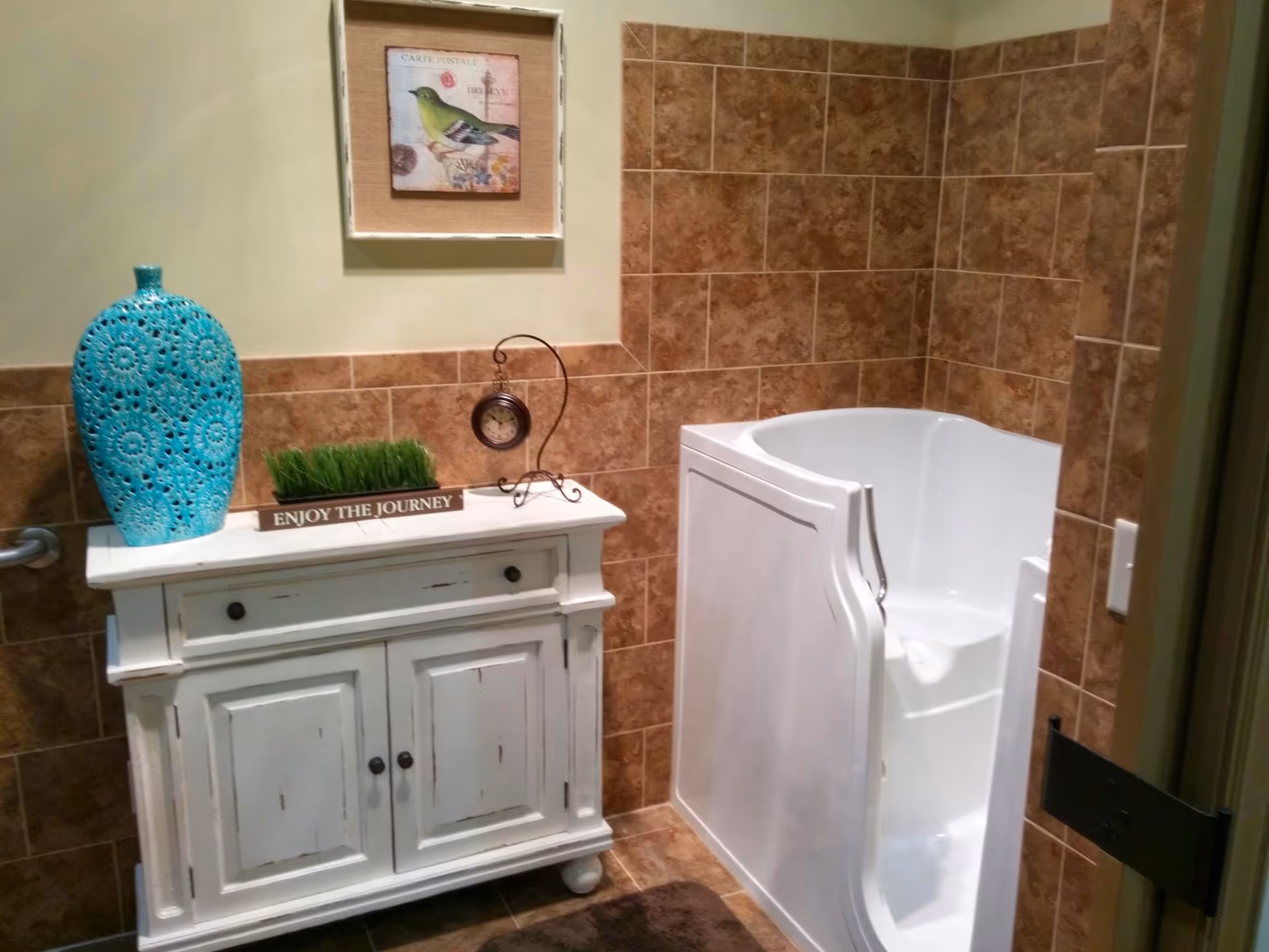 A bathroom with brown tiled walls and floor, featuring a white walk-in bathtub with a door. Next to the bathtub is a white distressed cabinet with a blue decorative vase, a small patch of artificial grass, and a small clock on top. Above the cabinet hangs a framed picture of a green bird.