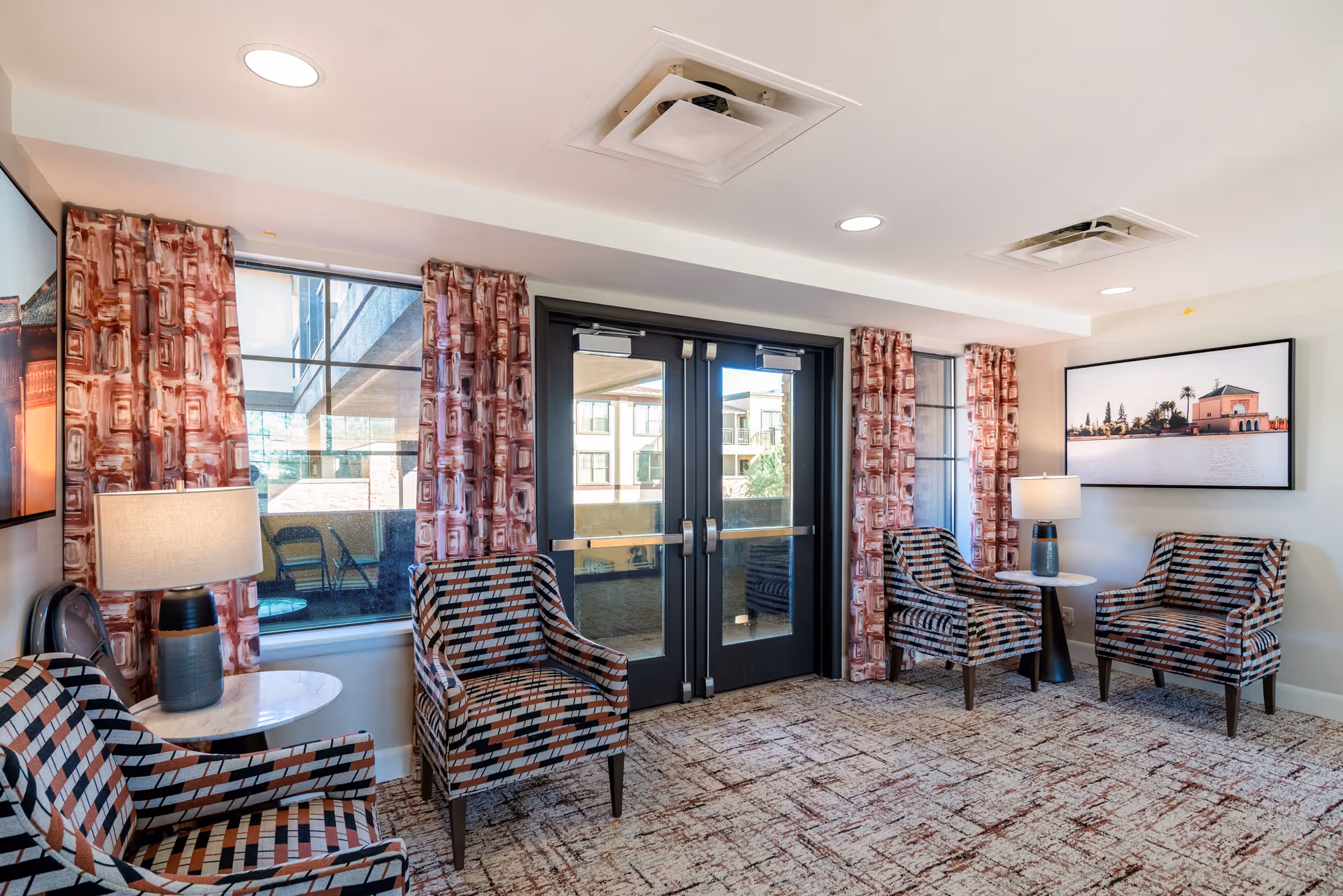 A bright and cozy sitting area with four patterned armchairs arranged around two small round tables with lamps. The room has large windows with patterned curtains and a glass door leading outside. A framed picture of a waterfront building hangs on the wall.