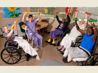 A group of elderly individuals and a caregiver participating in a seated exercise session indoors. The elderly participants, some in wheelchairs, are holding colorful exercise sticks above their heads and smiling. The room has a window with blinds, a round table, and a sunflower painting on the wall.