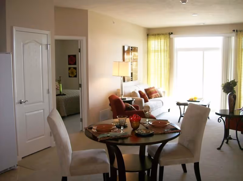 Open-plan living and dining area with a round set table and chairs in the foreground and a sofa and armchair by a large sunlit window with yellow curtains.