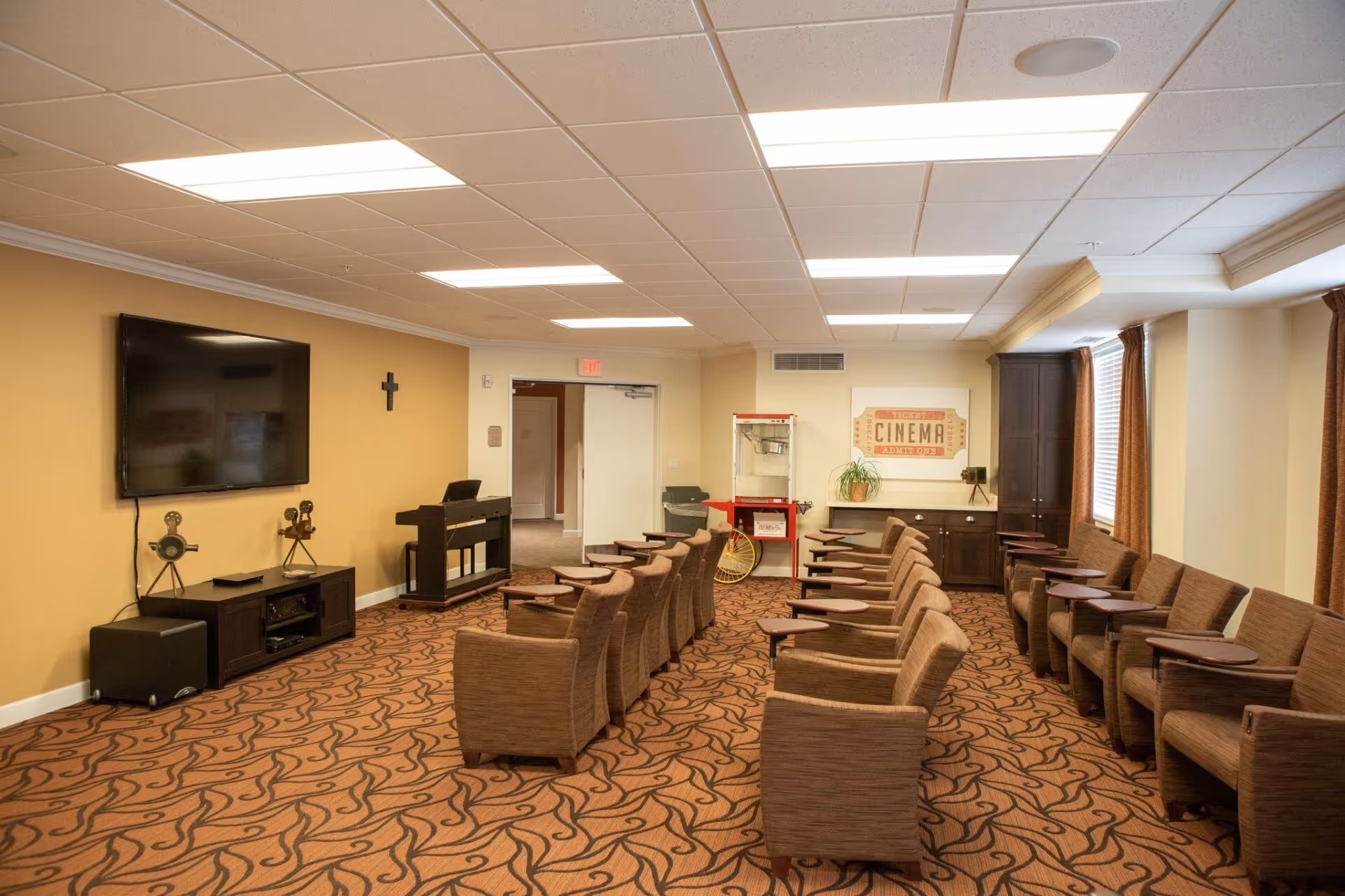 A cozy room with rows of brown upholstered chairs each with a small attached table, arranged facing a large flat-screen TV mounted on a beige wall. The room has patterned carpet, a popcorn machine, a piano, and a framed cinema ticket poster on the far wall. Windows with brown curtains allow natural light to enter.