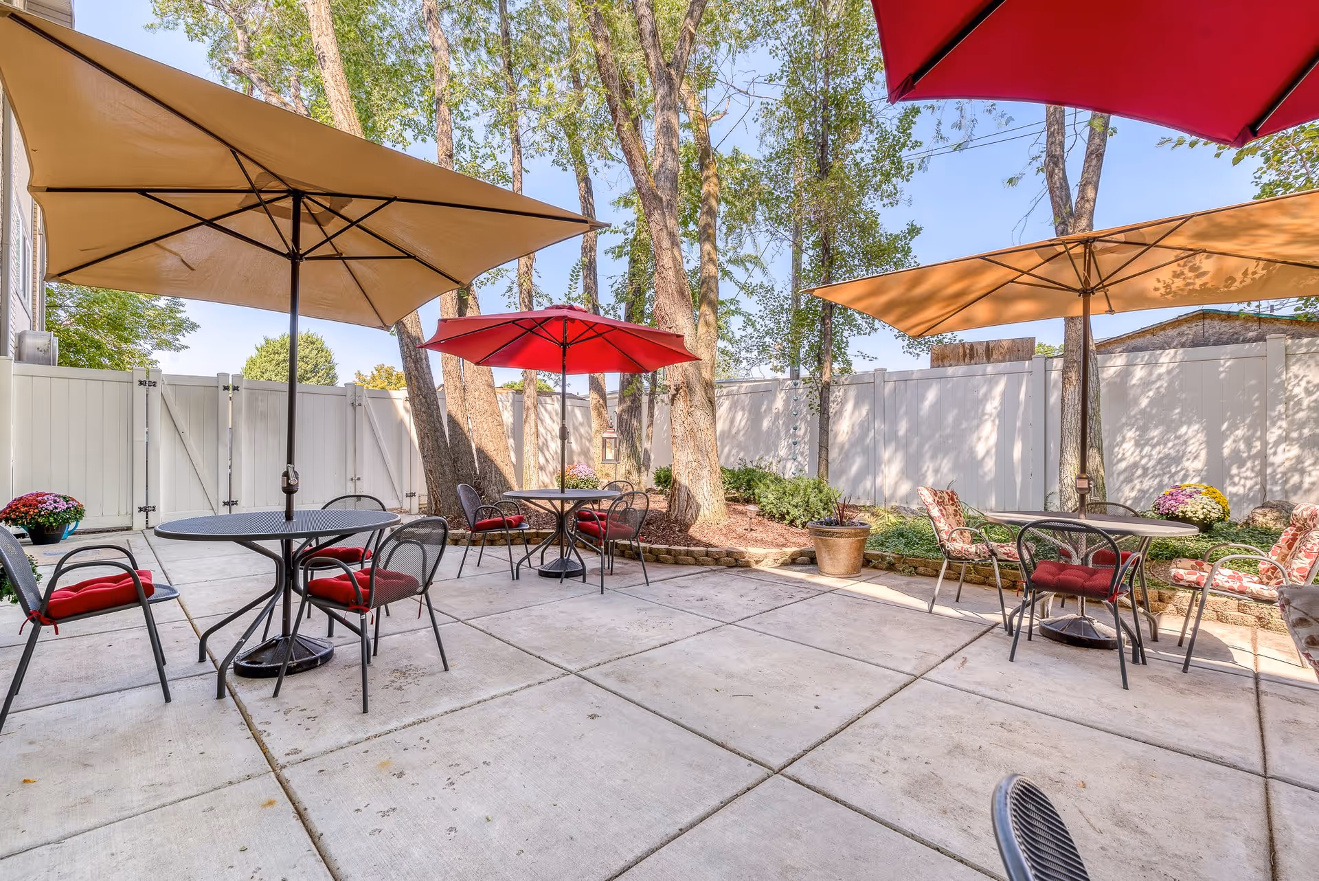 Outdoor patio area with several round metal tables and chairs, some with red cushions, shaded by large beige and red umbrellas. The patio is surrounded by a white fence and trees, with potted plants and landscaping along the edges.