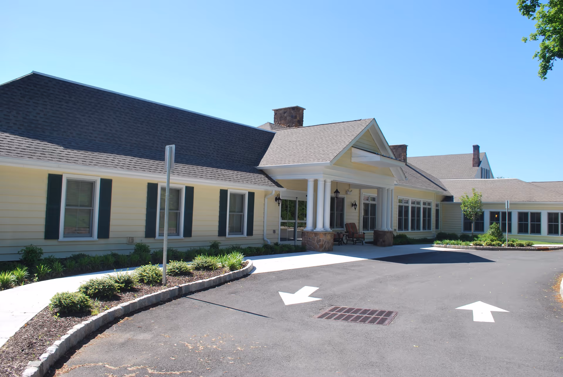 Exterior view of Canterbury Village facility showing a single-story building with a covered entrance supported by white columns, surrounded by landscaped bushes and a paved driveway with directional arrows.