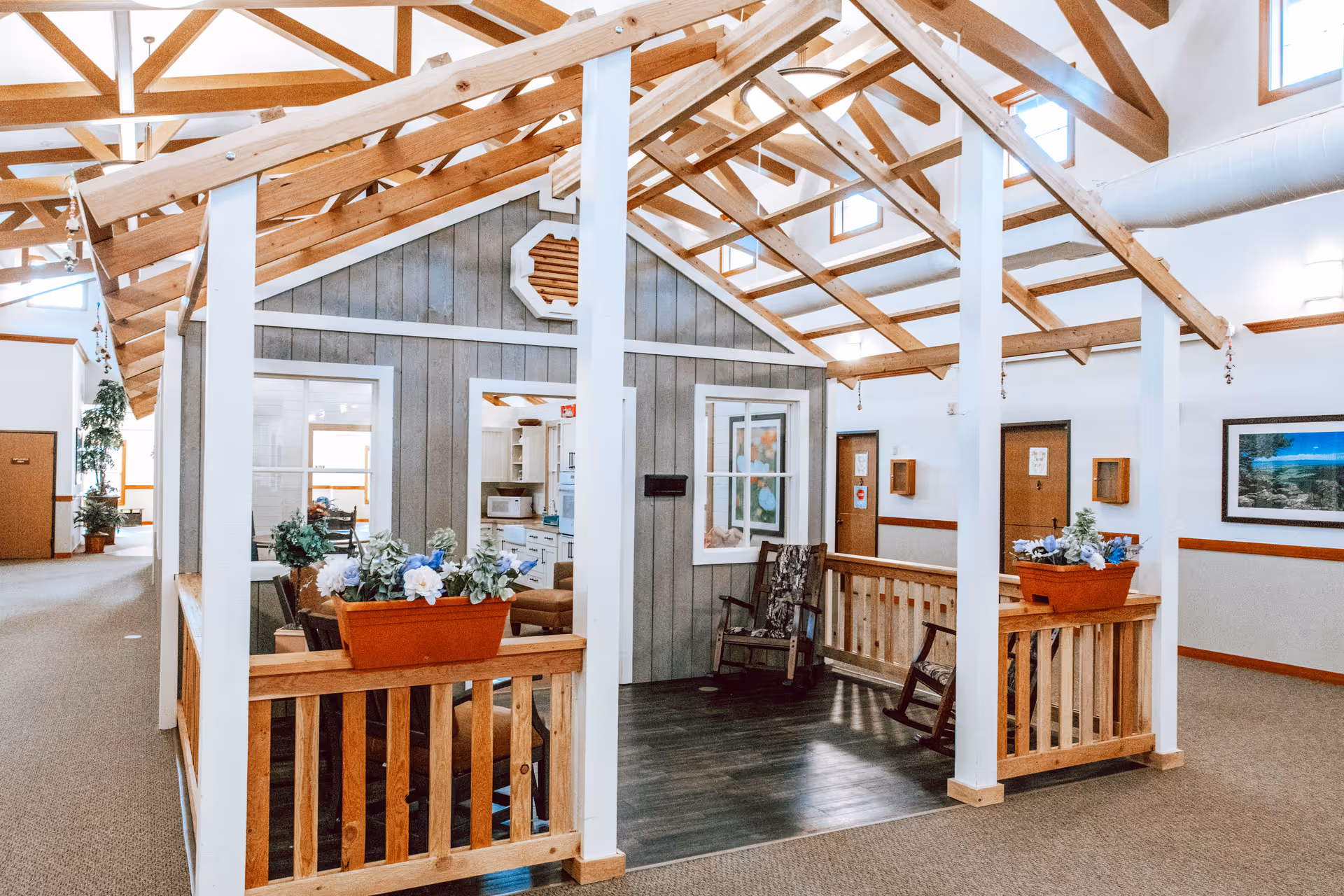 Interior view of a senior living facility featuring a small house-like structure with wooden beams and a pitched roof. The structure has gray paneling with white trim and contains rocking chairs and flower pots with artificial flowers. The surrounding area has carpeted floors, framed artwork on the walls, and several doors leading to other rooms.