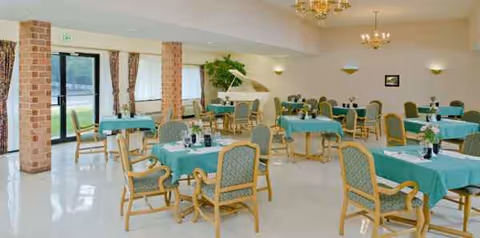 Bright dining hall with teal-covered tables, upholstered chairs, chandeliers, and large windows.