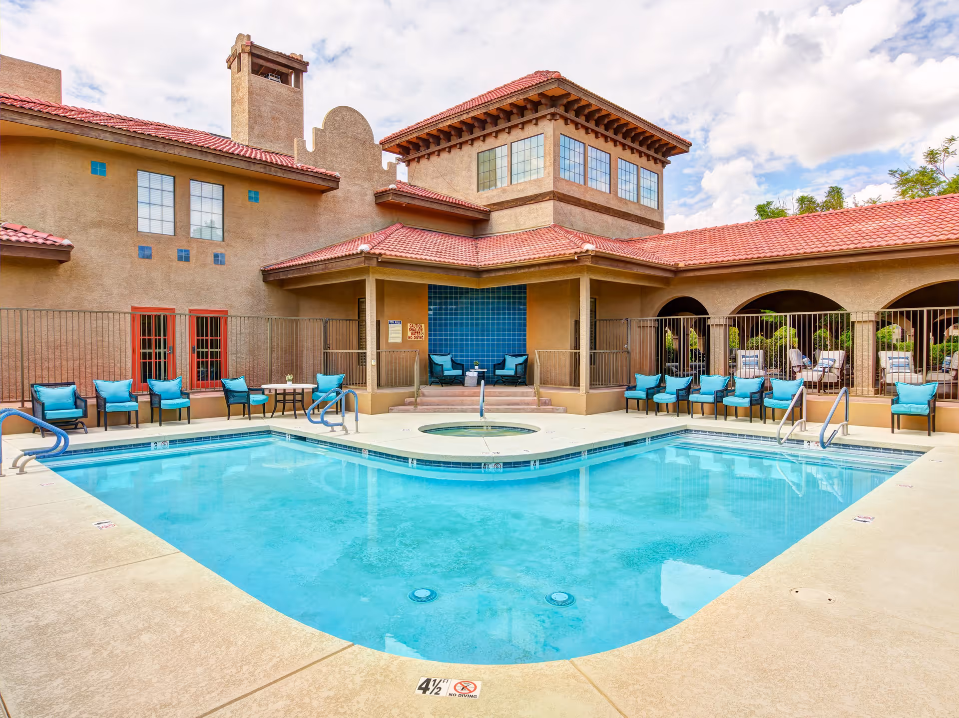 Outdoor swimming pool area at a senior living facility with turquoise water, surrounded by beige concrete deck and multiple blue cushioned chairs. The building has a tan stucco exterior with red tile roofing and large windows. There is a small hot tub adjacent to the pool and a covered seating area with two chairs and a small table.