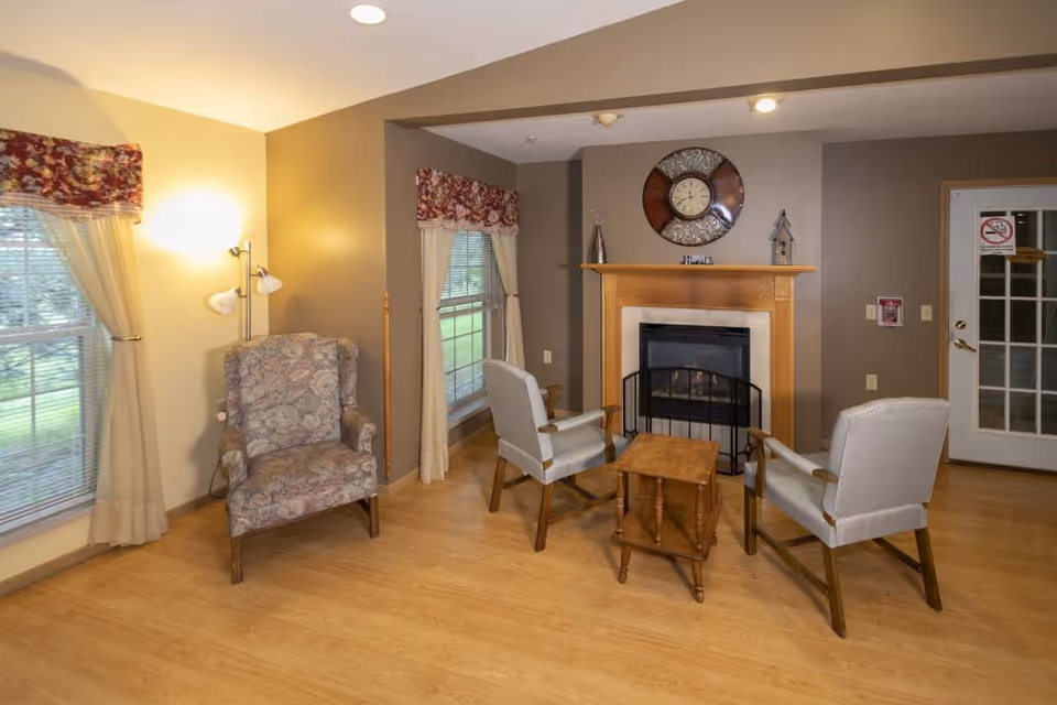A cozy living room area with a wooden floor, featuring a fireplace with a clock and decorative items on the mantel. There are three chairs arranged around a small wooden table, two upholstered armchairs and one floral patterned chair near a window with curtains and valances. The room has warm lighting and a door with glass panes leading outside.