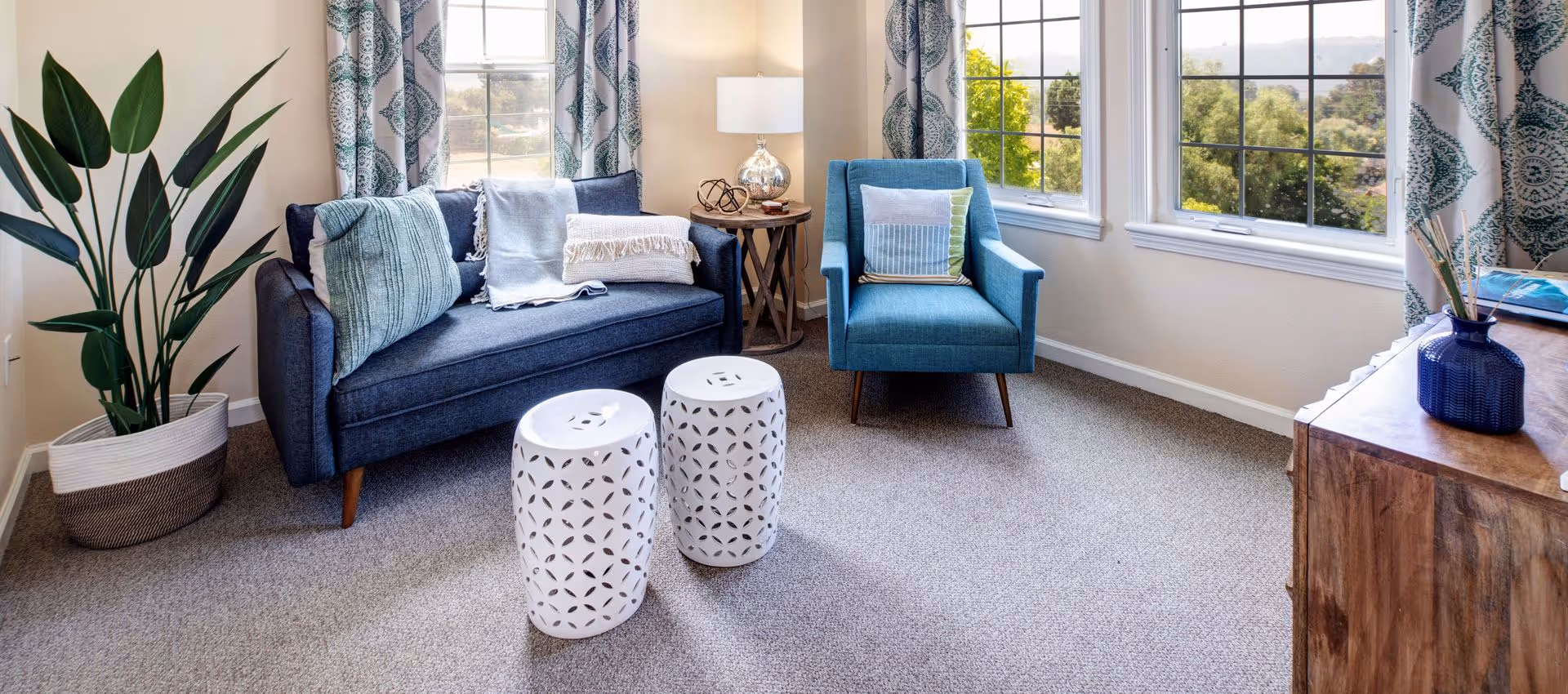Bright furnished living room with a navy sofa, teal armchair, patterned curtains, two white decorative stools, and large windows overlooking greenery.