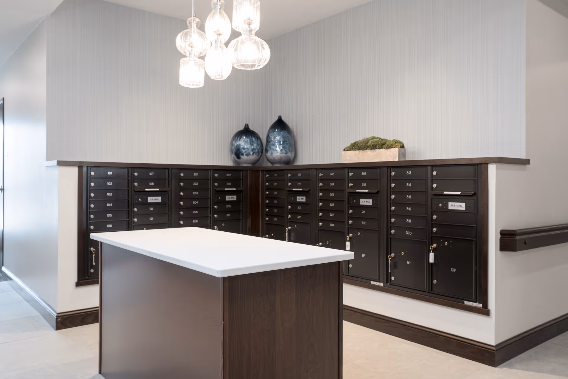 A bright mailroom interior with rows of dark resident mailboxes, a central white-topped island, hanging glass pendant lights, and decorative vases.