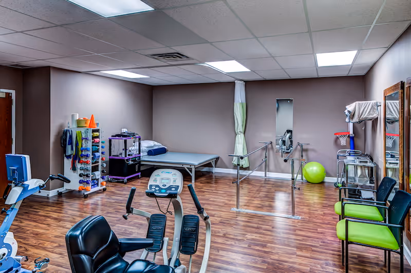 A senior living facility exercise and therapy room with wooden flooring, exercise equipment including a recumbent bike, parallel bars for walking support, a therapy table with a pillow, a rack with various exercise bands and weights, a green exercise ball, and chairs with green cushions along the wall. The room has beige walls and a drop ceiling with fluorescent lights.