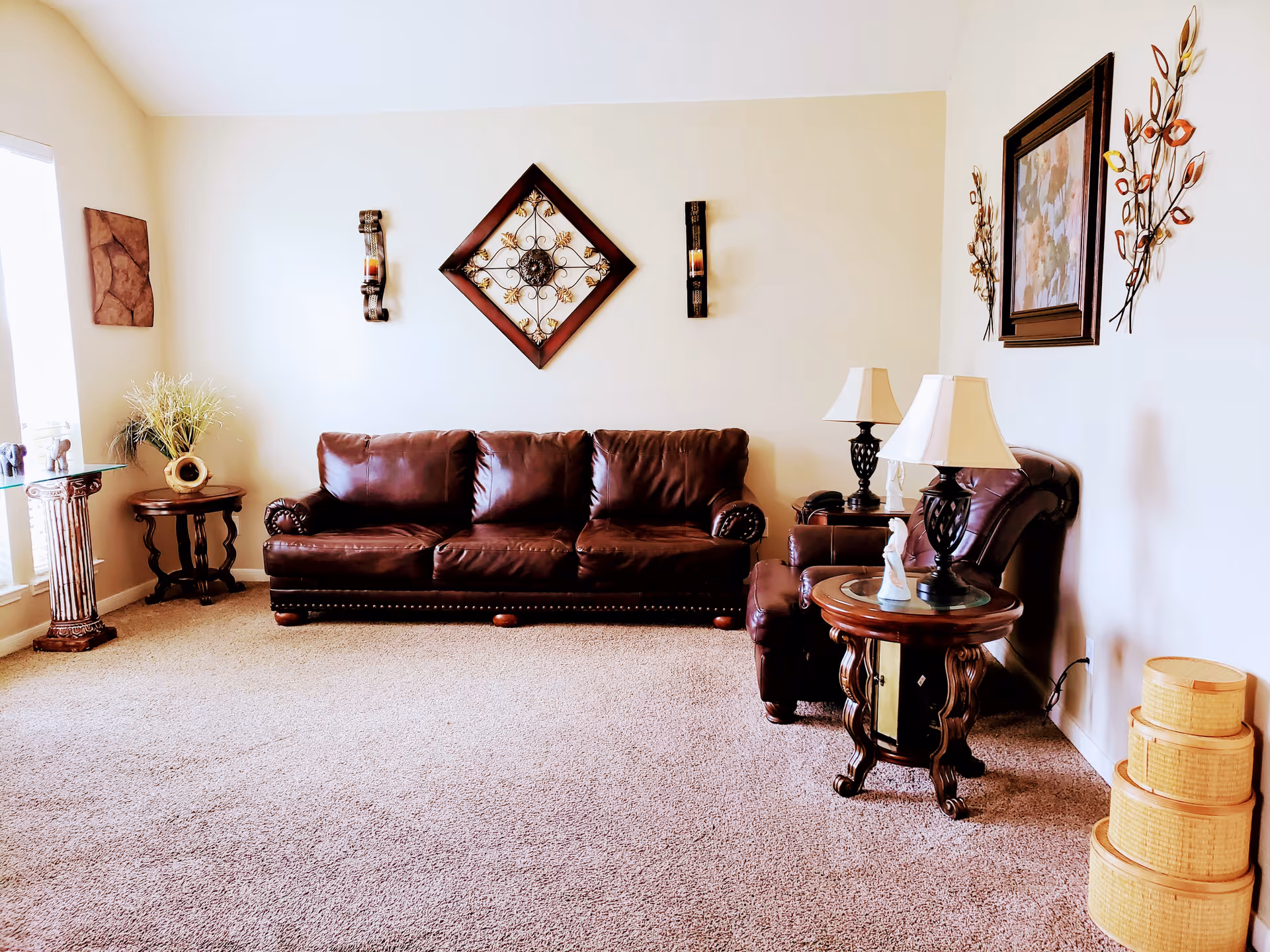 A cozy living room with a brown leather sofa and matching armchair. The walls are decorated with metal wall art and framed pictures. There are two side tables with lamps and decorative items, a small round table with a plant, and a set of three nested wicker baskets on the carpeted floor. Natural light comes in from a window on the left.