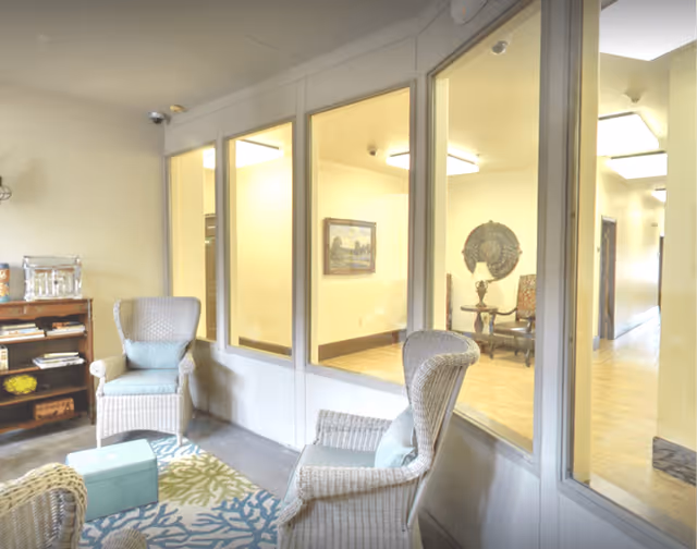 A cozy sitting area with wicker chairs and a small ottoman on a patterned rug, adjacent to a hallway visible through large interior windows. The hallway has wooden flooring and is decorated with framed artwork and a decorative wall piece.