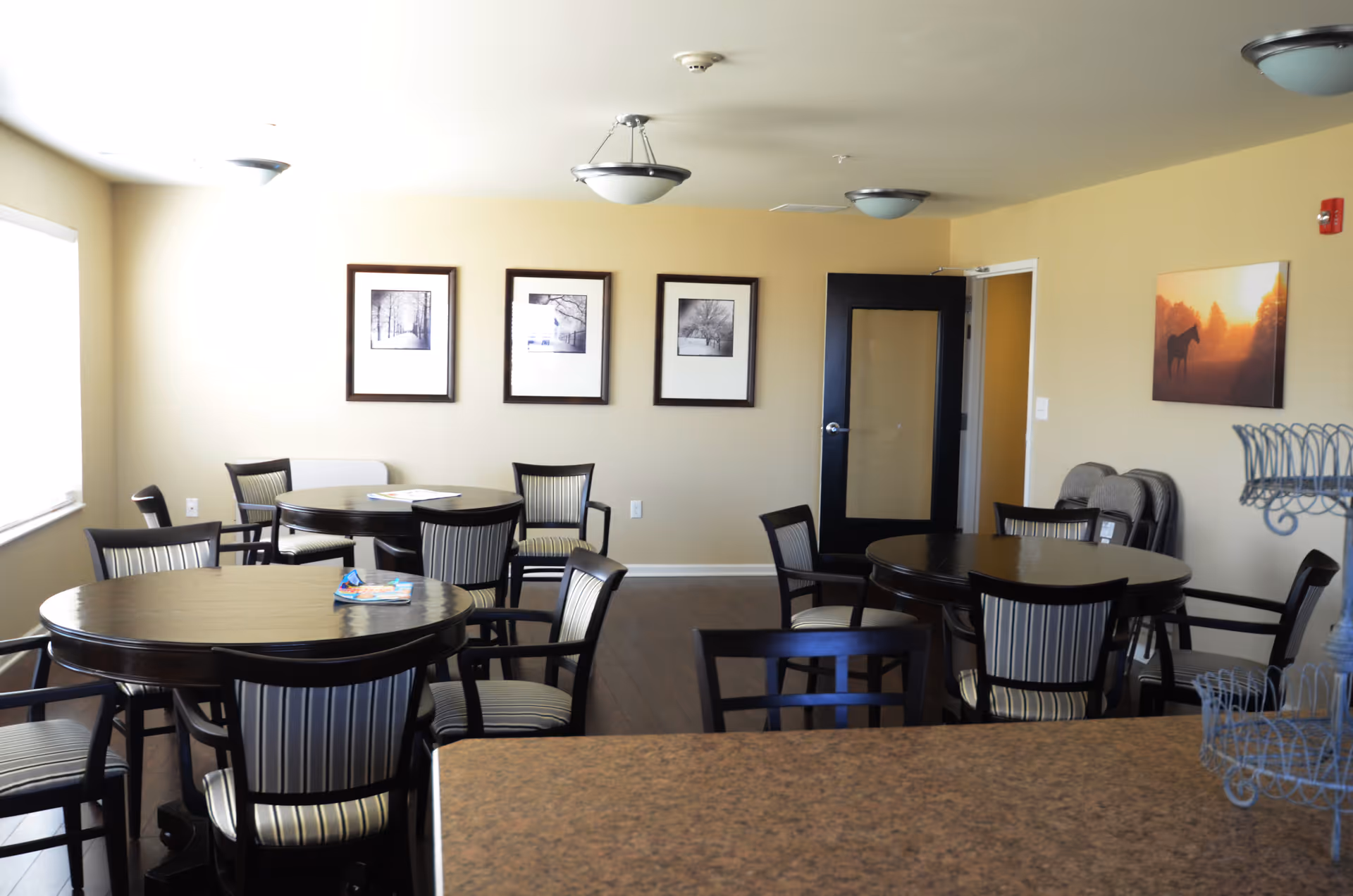 Bright common dining room with several round tables and striped chairs, framed artwork on the walls, and a black door.