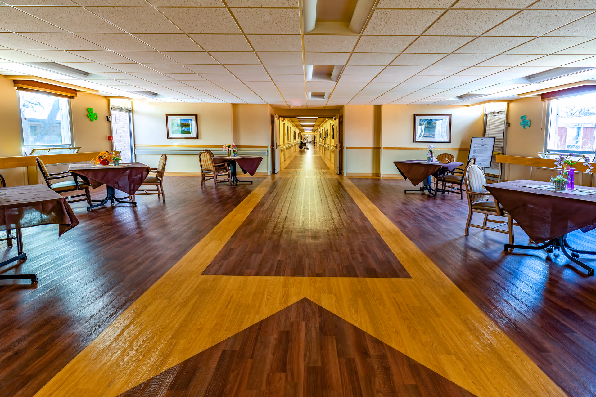 A spacious hallway in a senior living facility with a wooden floor featuring a large arrow design pointing down the corridor. On either side of the hallway, there are small tables covered with dark tablecloths and chairs, some with flower arrangements. The walls are light-colored with framed pictures and windows letting in natural light.
