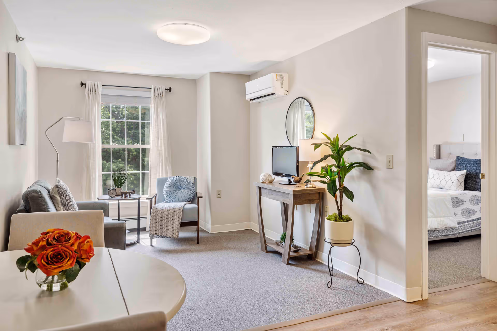 A bright and cozy living room area in a senior living facility with a gray sofa, a blue armchair, a floor lamp, and a small round table with a vase of orange roses. A large window with white curtains lets in natural light. A wooden console table with a small TV, a lamp, and decorative items is against the wall, next to a potted plant on a stand. An open doorway reveals a bedroom with a neatly made bed featuring white and blue bedding.