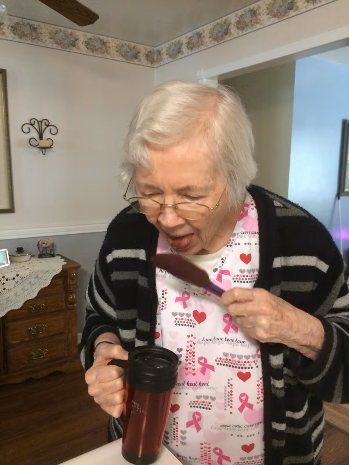 An elderly woman with white hair and glasses is standing indoors, holding a maroon travel mug in one hand and a purple spoon in the other. She is wearing a white shirt with pink ribbons and hearts, a black and gray striped cardigan, and appears to be about to eat or taste something from the spoon. The room has wooden furniture and floral wallpaper border near the ceiling.
