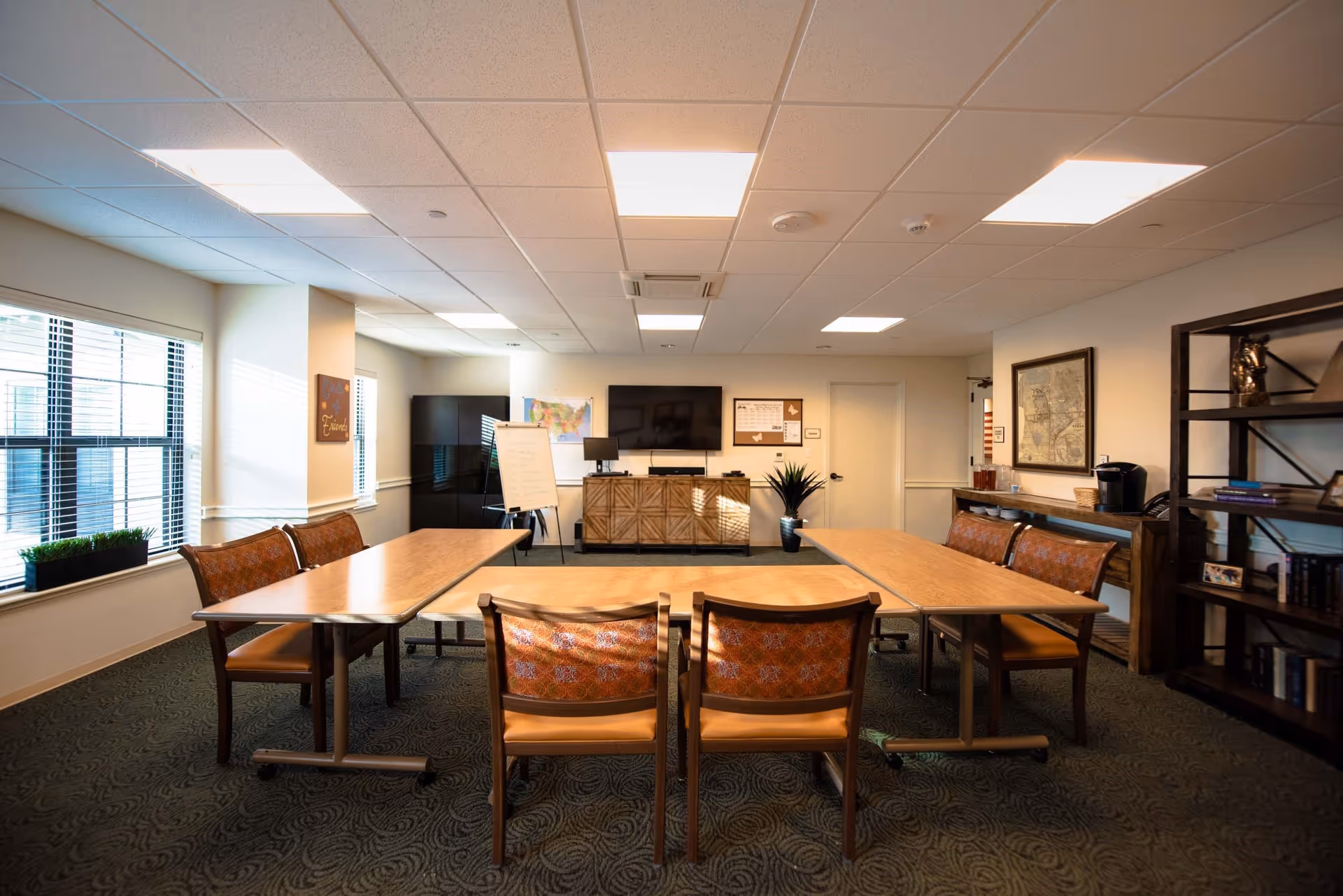 A well-lit meeting or activity room with tables arranged in a U-shape, surrounded by chairs with patterned upholstery. The room features large windows on the left side, a flat-screen TV mounted on the far wall, a whiteboard, a cabinet, a potted plant, and a bookshelf with books and decorative items. The carpet has a subtle pattern, and the ceiling has recessed lighting panels.