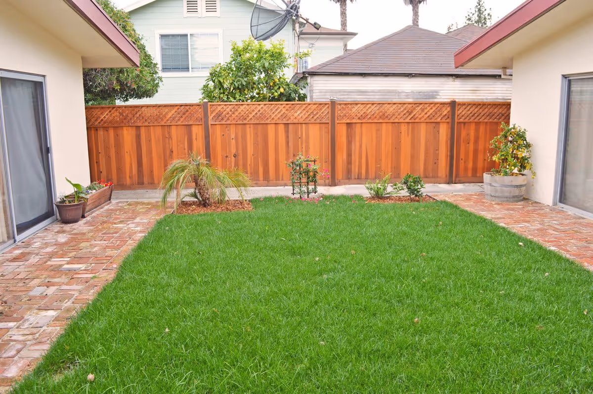 A small, well-maintained backyard garden area with a green lawn, bordered by brick pathways on both sides. There are a few small plants and shrubs planted in the lawn and pots along the edges. The area is enclosed by a wooden fence, with neighboring houses visible in the background.