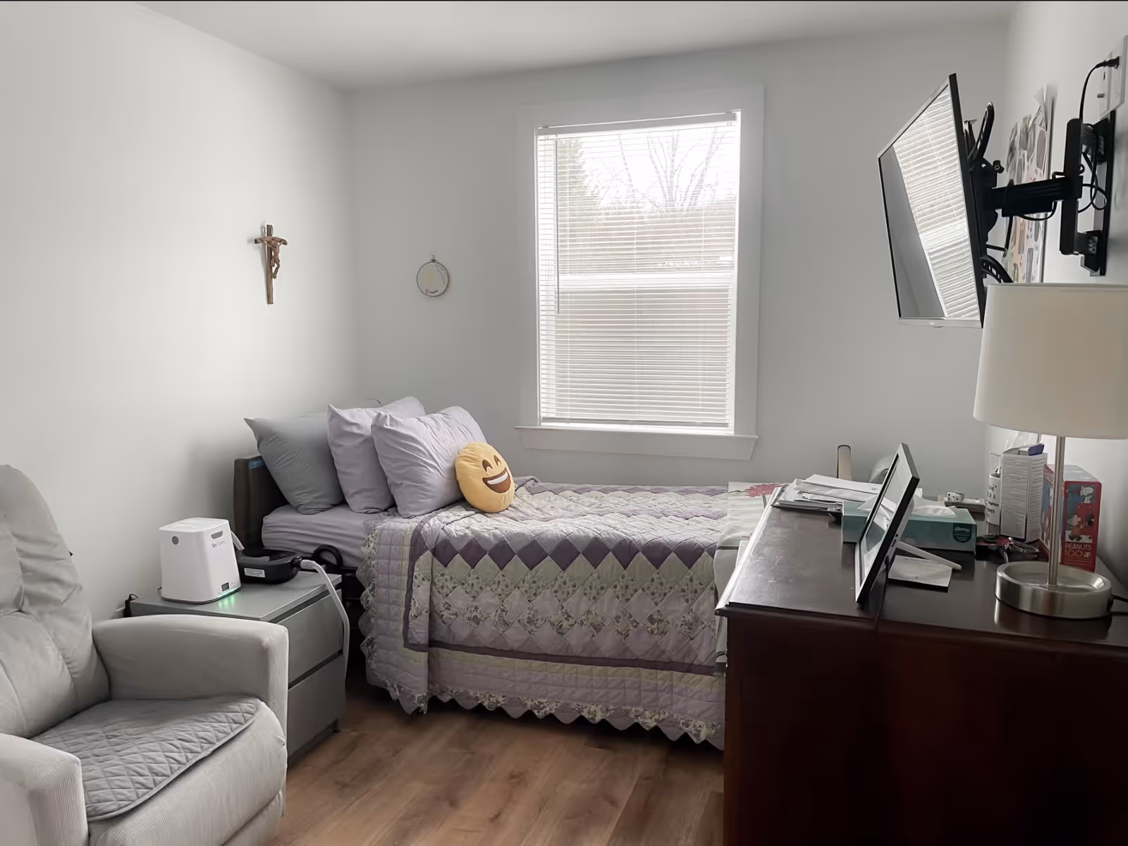 A small, well-lit bedroom with a single bed covered by a purple quilt and emoji pillow, an armchair, dresser with lamp and a wall-mounted TV.