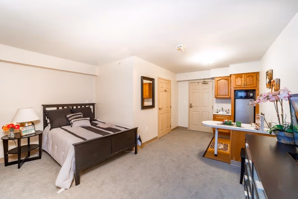 A studio apartment interior with a bed on the left side, a nightstand with a lamp and flowers, a small kitchen area with wooden cabinets, a refrigerator, a sink, and a small counter with two green cups. There is a door in the background and a mirror on the wall. The room has beige walls and carpeted floor.