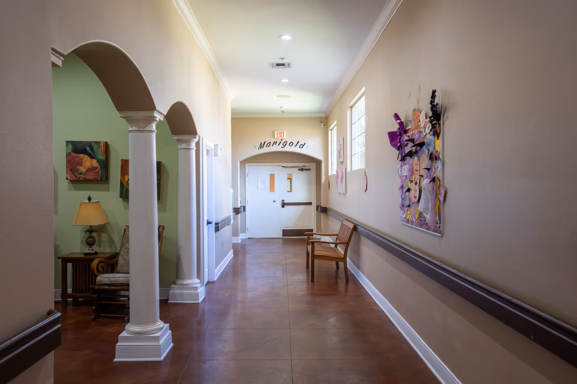 A well-lit hallway in a senior living facility with beige walls and brown flooring. The hallway features white columns on the left side leading to a sitting area with a lamp and chair. On the right side, there is a wooden bench and colorful artwork displayed on the wall. At the end of the hallway, double doors are visible with a sign above reading 'Marigold'.