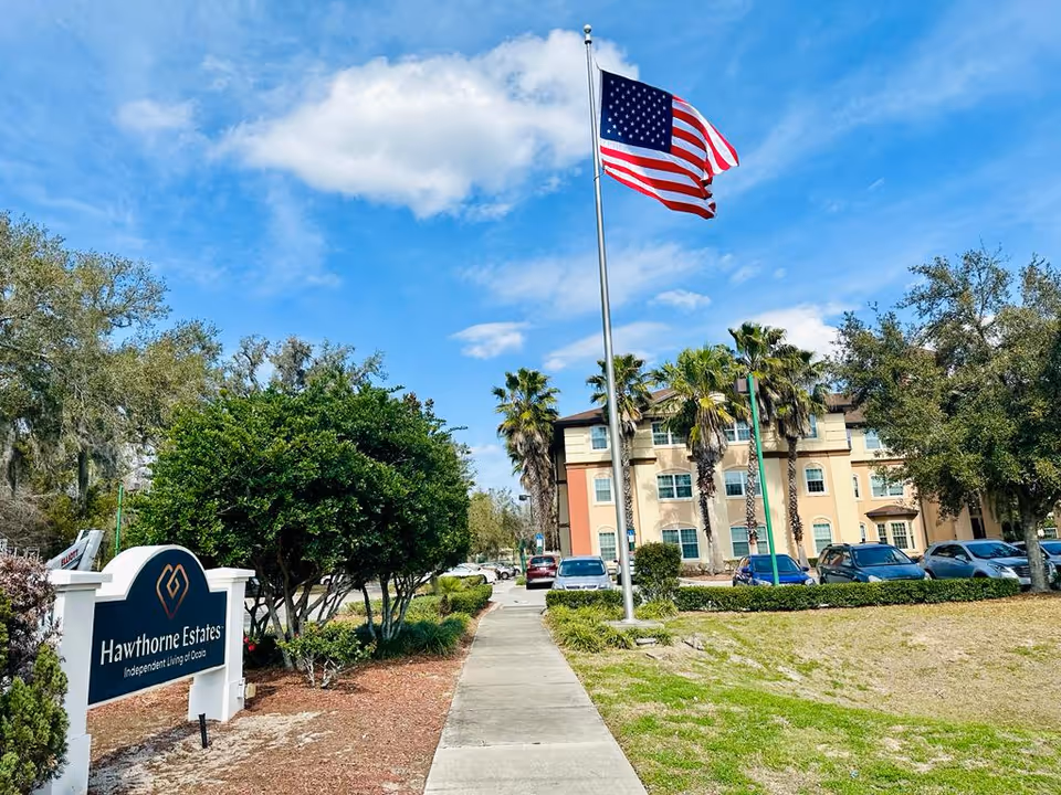 Entrance walkway to Hawthorne Estates with a sign, a tall American flag, and the facility building and parked cars in the background.