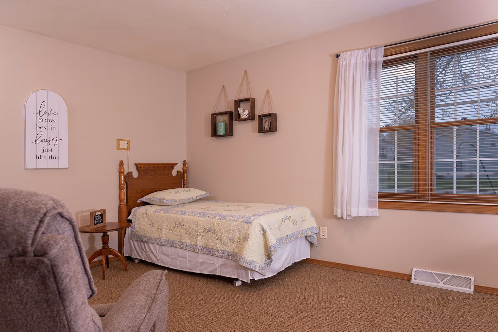 A tidy single bedroom with a twin bed, wooden headboard, nightstand, armchair, wall decor, and a window with blinds and curtains.