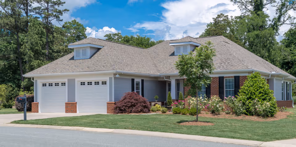 A single-story residential building with a gray roof, light gray siding, and brick accents. The house features two white garage doors, black window shutters, and a well-maintained front yard with green grass, bushes, and a small tree. The sky is partly cloudy with blue patches visible.