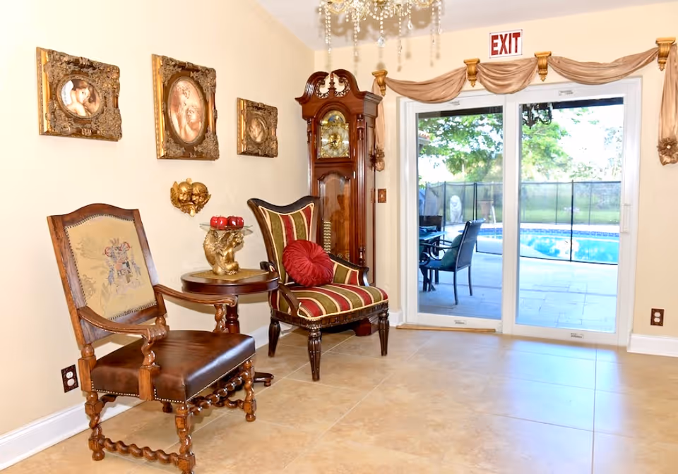 A cozy interior corner with two antique-style chairs, a small round table with a decorative golden sculpture holding red apples, and a tall grandfather clock. The wall is adorned with three framed vintage portraits. A sliding glass door with an exit sign above it leads to an outdoor patio with a swimming pool and patio furniture visible outside.