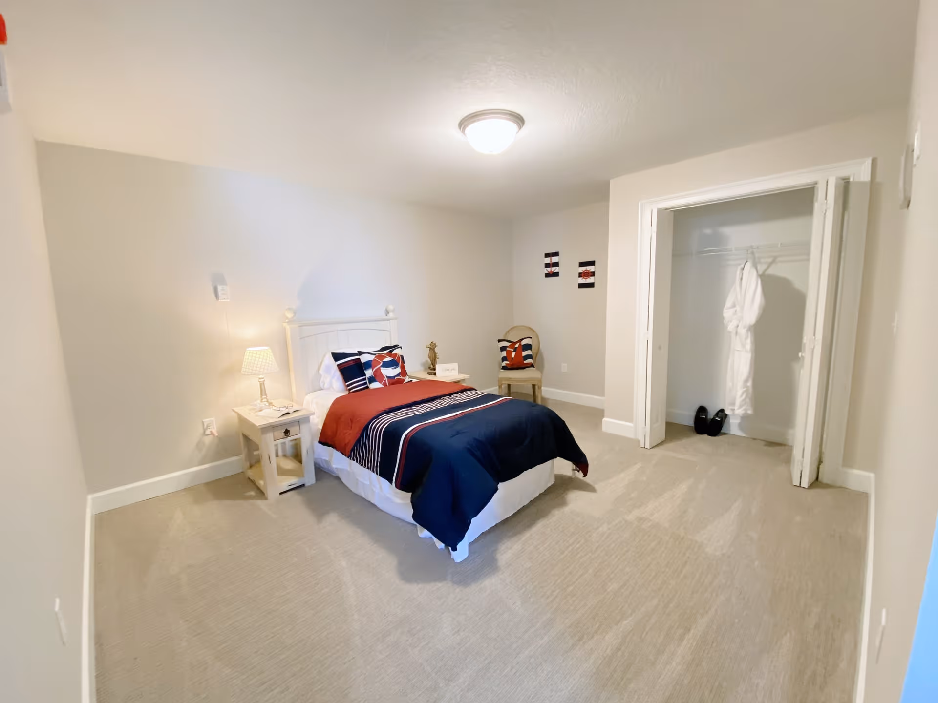 A neatly arranged bedroom with a single bed dressed in navy blue, red, and white bedding. There is a small white nightstand with a lamp and a book beside the bed. A chair with a decorative pillow is placed near an open closet that contains a white robe and a pair of black shoes. The room has beige carpet and light-colored walls with two small framed artworks.