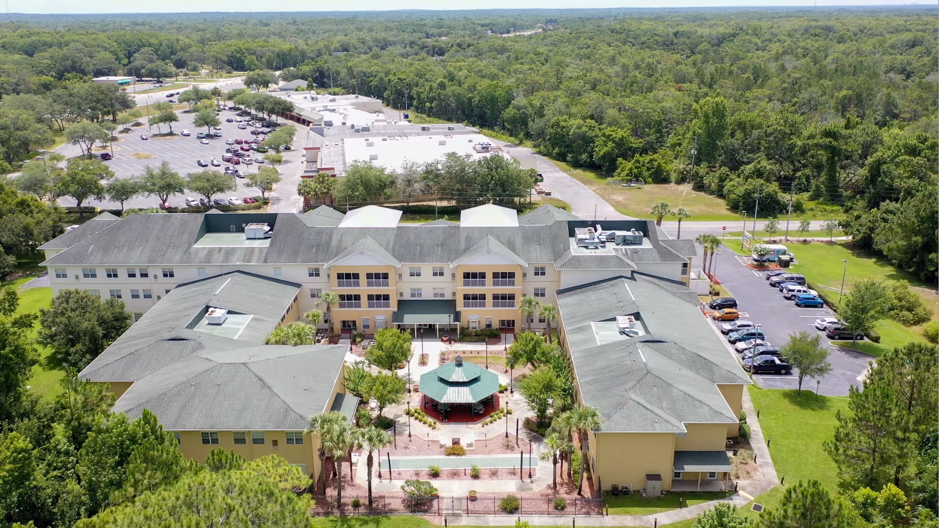 Aerial view of Sunflower Springs senior living facility showing a large, multi-wing building with a central courtyard featuring a gazebo. Surrounding the building are green lawns, trees, and parking lots with several cars. The background includes a dense forested area under a clear sky.