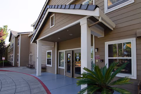 Exterior view of the entrance to Vista Roseville Senior Living facility showing a covered porch with double glass doors, beige siding, windows, and a small palm plant near the entrance.