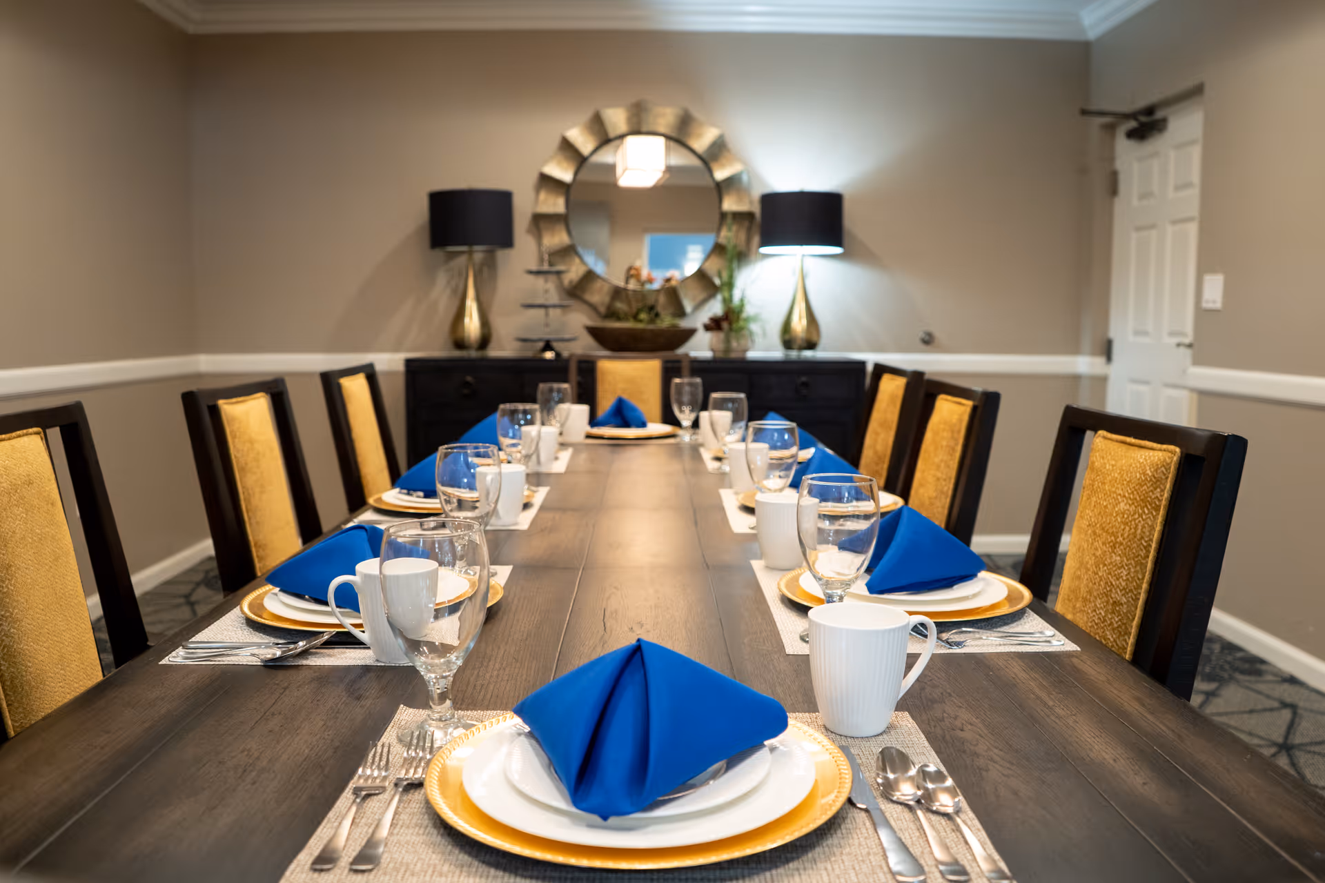 A long dining table set for a meal with blue folded napkins, white plates on gold chargers, white mugs, and clear water glasses. The table is surrounded by chairs with yellow upholstery. In the background, there is a sideboard with two gold lamps with black shades and a decorative round mirror on the wall.