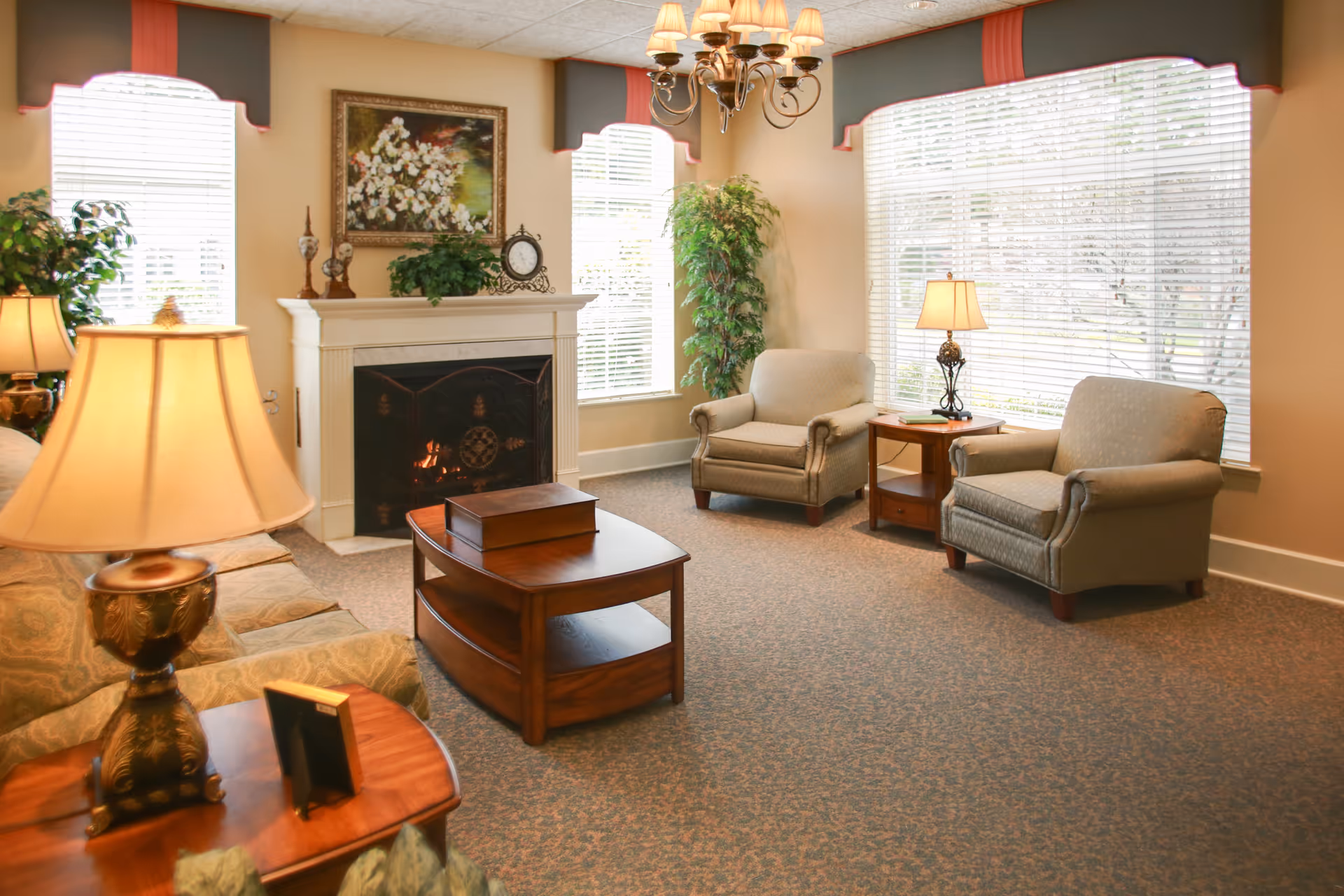 A cozy living room with a lit fireplace, two armchairs, a sofa, wooden coffee and side tables, table lamps, a chandelier, large windows with blinds, and decorative plants and artwork.