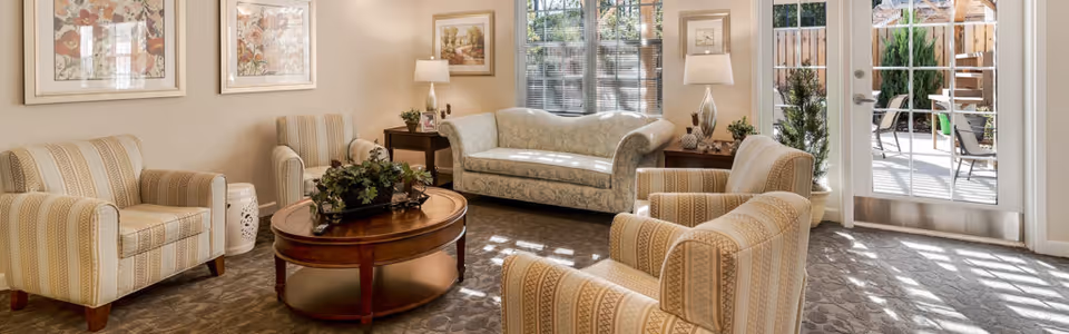 A bright and cozy living room with a patterned sofa, two striped armchairs, a round wooden coffee table with a plant centerpiece, two side tables with lamps, framed artwork on the walls, and large windows and glass doors letting in natural light and showing an outdoor patio area.