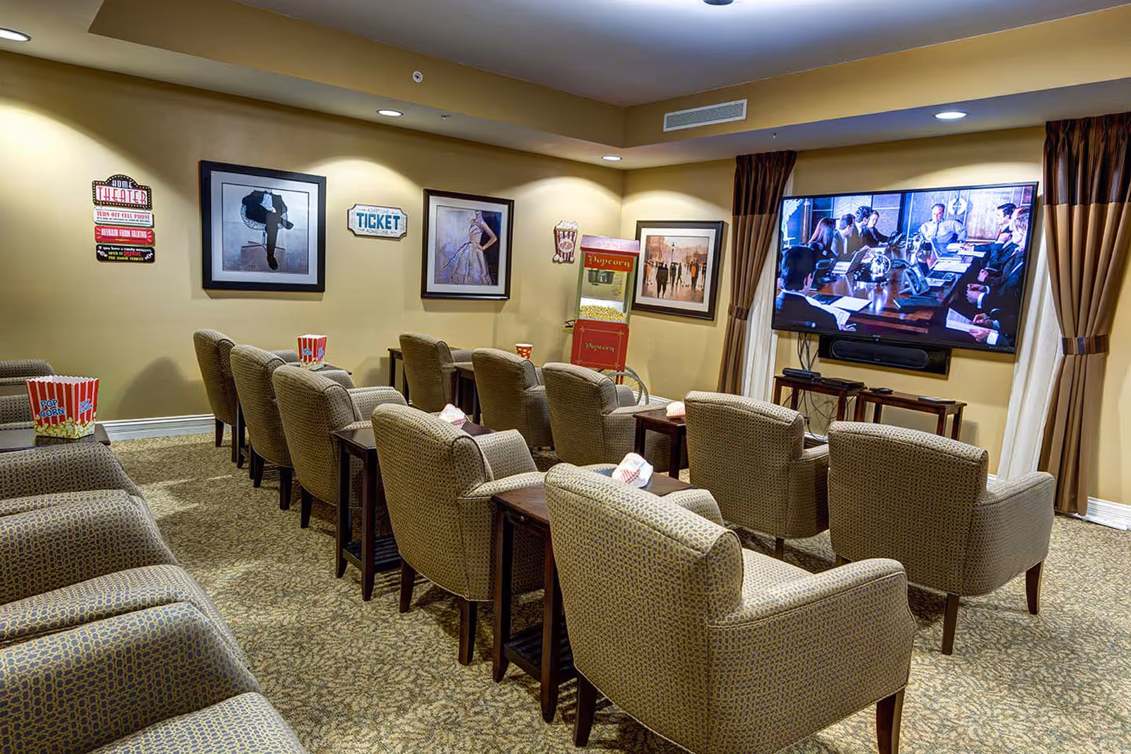 A cozy community movie room with rows of upholstered chairs facing a large wall-mounted TV, popcorn boxes, and movie-themed decor.