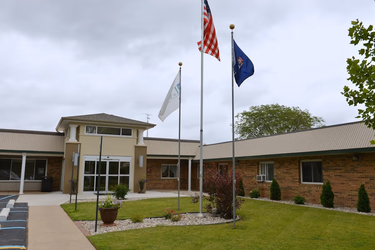Exterior view of a single-story brick building with a beige entrance featuring glass doors. Three flagpoles with flags, including the American flag, stand in front of the building. There is a well-maintained lawn with small bushes and a paved walkway leading to the entrance under a cloudy sky.