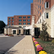 Exterior view of the entrance to a multi-story senior living facility with a covered drop-off area, a paved driveway, and a landscaped garden with colorful flowers along the walkway.