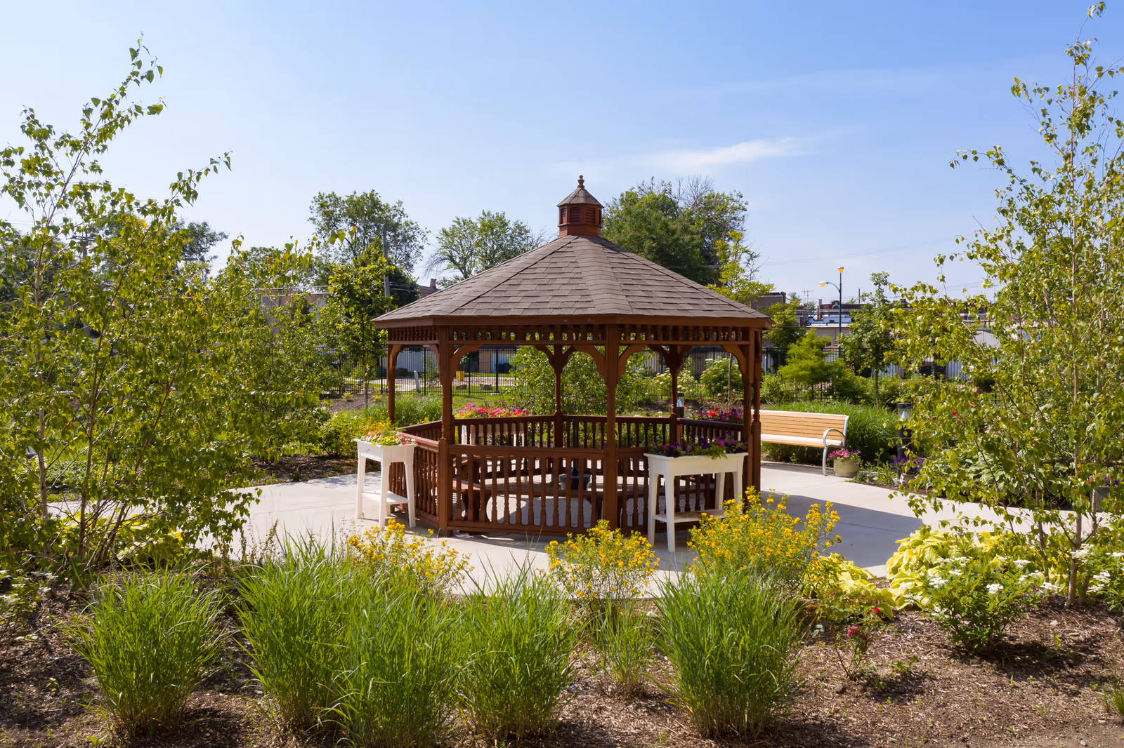 A wooden gazebo with a shingled roof is situated in a garden area surrounded by green shrubs, yellow flowers, and trees under a clear blue sky. There are benches and small tables with flower pots around the gazebo.