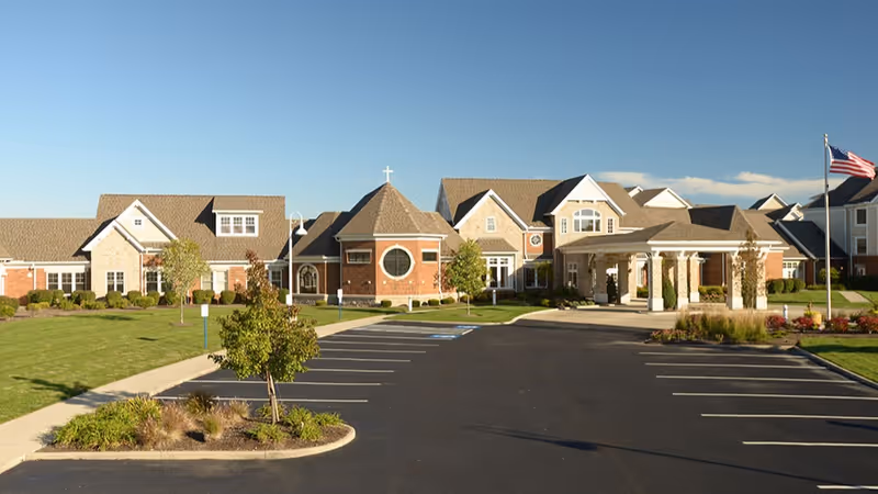 Exterior view of Saint Therese at St. Mary of the Woods facility showing a large building with a mix of brick and siding, multiple roof peaks, a circular window with a cross on top, a covered entrance, landscaped grounds, and an empty parking lot with an American flag on a flagpole.