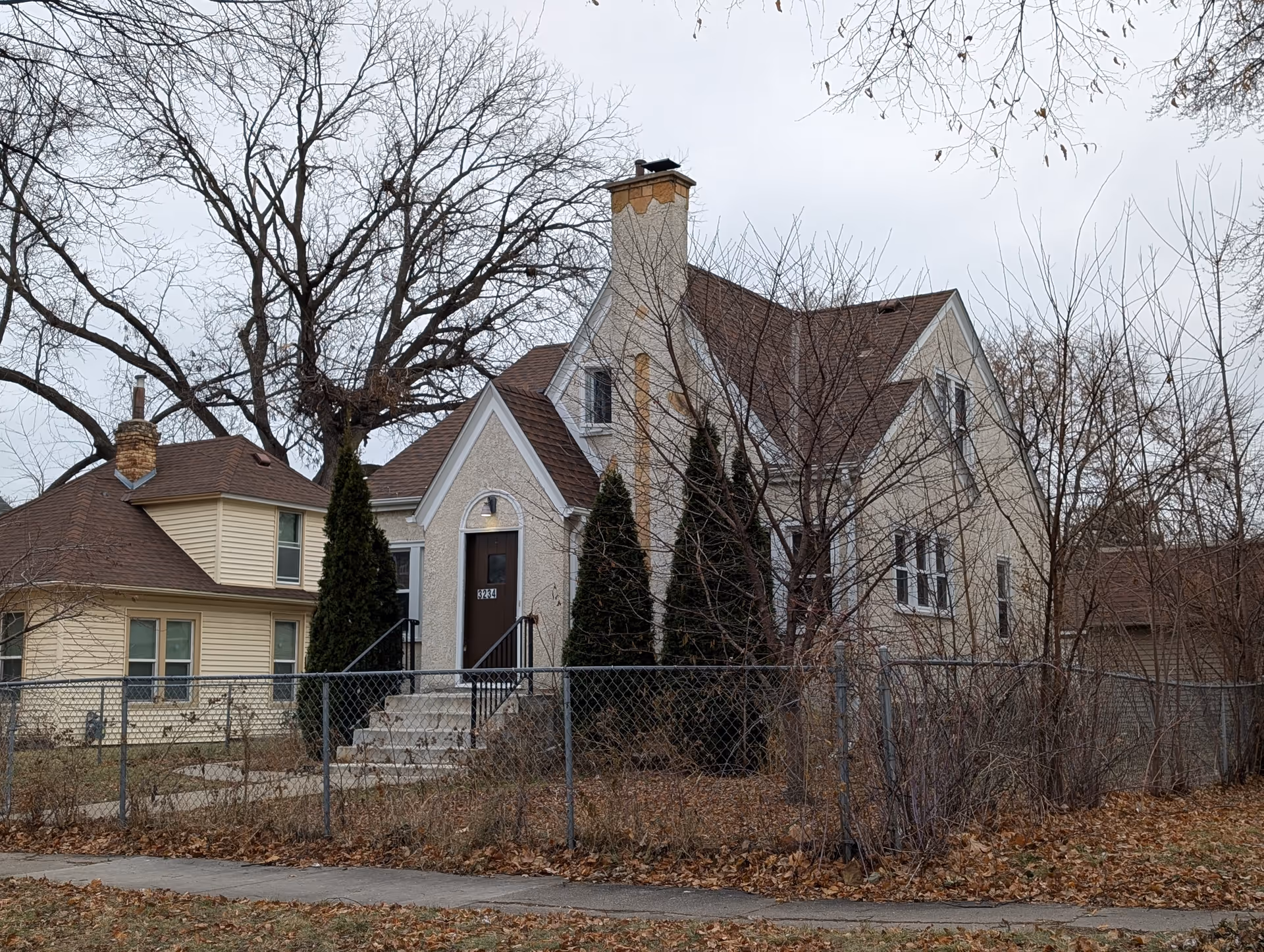 Front exterior of a two-story house with a steep roof and chimney, front steps leading to a door, a chain-link fence, and leafless trees.