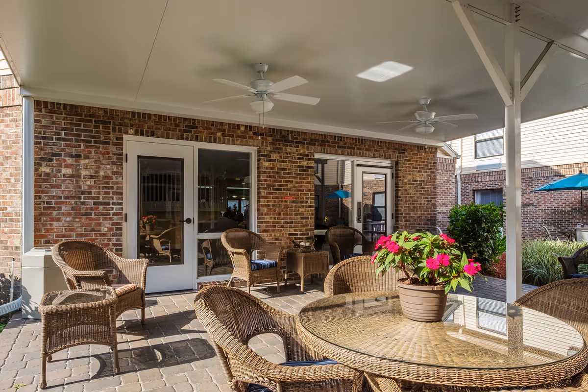 Covered outdoor patio area with wicker chairs and a glass-top table with a potted plant featuring pink flowers. The patio has a brick wall with two glass doors and ceiling fans overhead.
