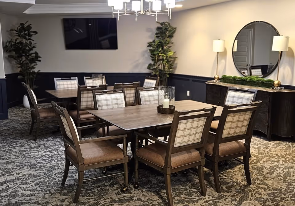 A dining room with two wooden tables surrounded by cushioned chairs with plaid backs. The room has patterned carpet flooring, a large round mirror on the wall above a dark wooden sideboard, two table lamps, and two tall potted plants. A flat-screen TV is mounted on the wall.