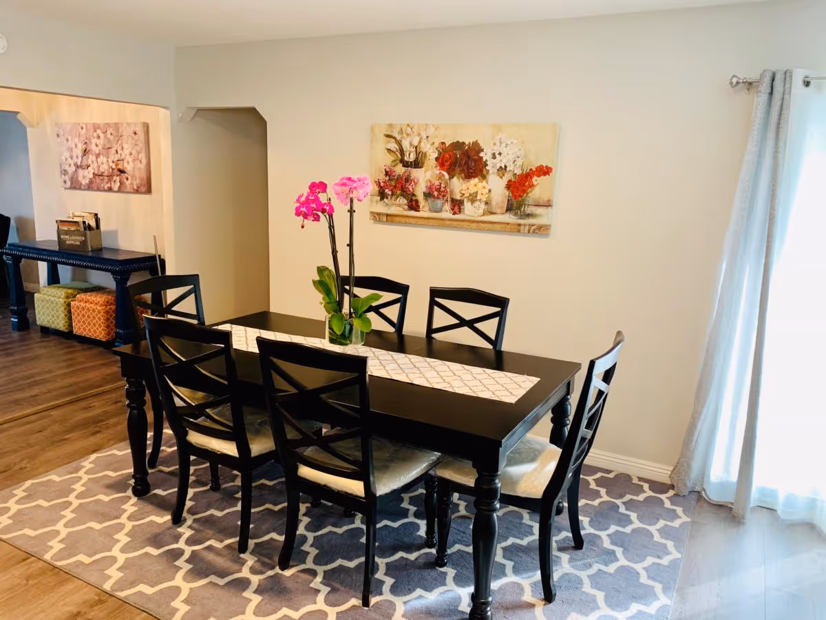 A dining room with a black rectangular table and six matching chairs. A white and gray patterned rug is underneath the table. A vase with pink flowers is centered on a white table runner. The walls are light-colored with a floral painting hanging above the table. To the right, light gray curtains cover a window or glass door, and to the left, there is an open doorway leading to another room with a console table and colorful ottomans underneath.